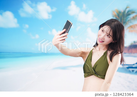 快晴のビーチでスマホで自撮りをする水着を着た若い女性 128423364