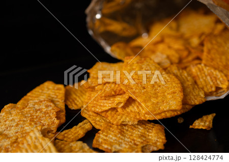 Golden crunchy ridged potato chips spilling from a bag onto a dark background, symbolizing delicious indulgent snack or unhealthy food choice Golden crunchy ridged potato chips spilling from a bag onto a dark background, symbolizing delicious indulgent snack or unhealthy food choice 128424774