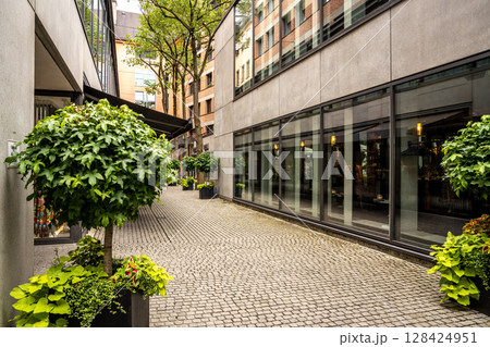 Modern Street Scene In Munich Center: Contemporary Architecture With Glass Facades And Greenery In A Quiet Urban Courtyard 128424951