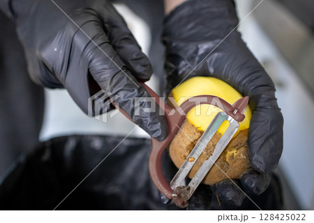 Person's hands wearing black safety gloves expertly peeling a fresh brown potato with a kitchen peeler, emphasizing food preparation and culinary hygiene Person's hands wearing black safety gloves expertly peeling a fresh brown potato with a kitchen peeler, emphasizing food preparation and culinary hygiene 128425022