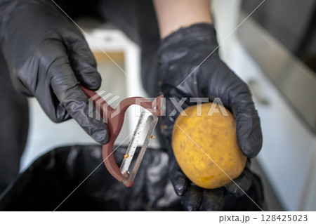 Hands in black protective gloves peeling a fresh raw potato using a kitchen peeler, highlighting essential food safety and hygiene for culinary preparation 128425023