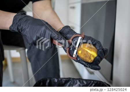 Person's gloved hands meticulously peeling a fresh raw potato with a sharp vegetable peeler, emphasizing hygienic food preparation in a contemporary kitchen environment 128425024