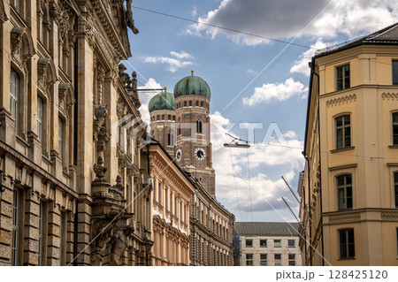 Iconic Cathedral Of Our Dear Lady In Munich: Majestic Twin Domes Rising Above Historic Buildings Under A Bright Blue Sky With Scenic Clouds Near Marienplatz 128425120