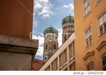Iconic Cathedral Of Our Dear Lady In Munich: Majestic Twin Domes Rising Above Historic Buildings Under A Bright Blue Sky With Scenic Clouds Near Marienplatz Iconic Cathedral Of Our Dear Lady In Munich: Majestic Twin Domes Rising Above Historic Buildings Under A Bright Blue Sky With Scenic Clouds Near Marienplatz 128425229