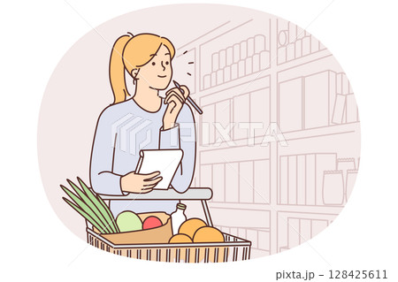 Woman walks around supermarket with cart filled with vegetables and holds checklist in hands 128425611