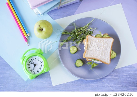 On the table is a school lunch, an alarm clock and stationery. A sandwich, an apple and a bottle of water. School lunch and textbooks. On the table is a school lunch, an alarm clock and stationery. A sandwich, an apple and a bottle of water. School lunch and textbooks. 128426210