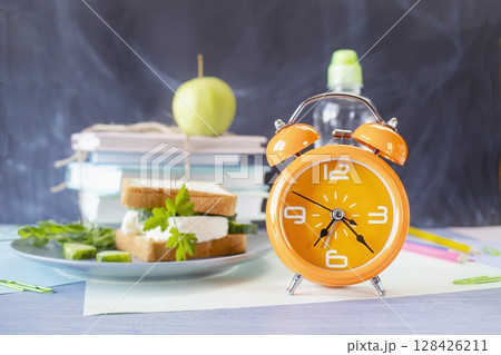 On the table is a school lunch, an alarm clock and stationery. A sandwich, an apple and a bottle of water. School lunch and textbooks. On the table is a school lunch, an alarm clock and stationery. A sandwich, an apple and a bottle of water. School lunch and textbooks. 128426211