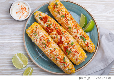 Elotes mexican street corn cob covered in spices and cheese being served on plate Elotes mexican street corn cob covered in spices and cheese being served on plate 128426556