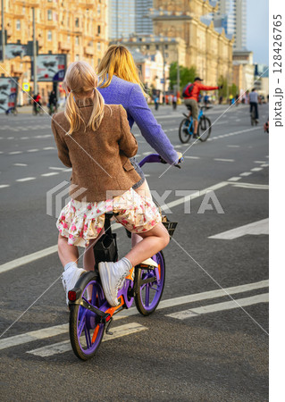 Two women riding one bicycle through city street during public cycling event. Concept of friendship, joy, freedom in motion 128426765