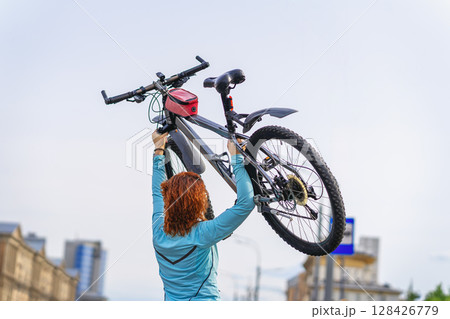 Woman holding bicycle triumphantly above head against clear sky after urban ride, celebrating success and power. Concept of cycling, freedom, cycling, personal sport achievement outdoors Woman holding bicycle triumphantly above head against clear sky after urban ride, celebrating success and power. Concept of cycling, freedom, cycling, personal sport achievement outdoors 128426779