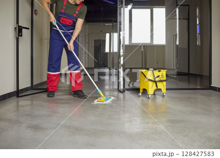 Janitor in uniform mopping floor inside modern commercial building during routine cleaning shift. 128427583