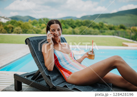 Young woman in swimsuit sitting by pool, making phone call. 128428196
