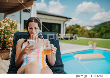 Young woman sitting by swimming pool, scrolling on smartphone. Young woman sitting by swimming pool, scrolling on smartphone. 128428197