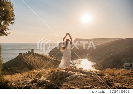 woman stands on a rocky hill overlooking a body of water. She is wearing a white dress and she is in a state of joy or celebration. Concept of freedom and happiness. woman stands on a rocky hill overlooking a body of water. She is wearing a white dress and she is in a state of joy or celebration. Concept of freedom and happiness. 128428491