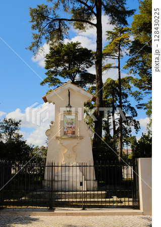 Catholic roadside shrine in Fatima Portugal with tiled Virgin Mary image and metal fence surrounded by pine trees under clear blue sky religious monument and local devotion site. High quality photo 128430225