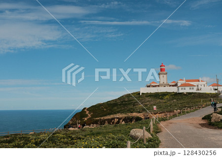 Lush green hills and rugged terrain with blue sky and scattered clouds in Cabo da Roca Sintra Portugal natural landscape with vegetation from scenic hiking trail Lush green hills and rugged terrain with blue sky and scattered clouds in Cabo da Roca Sintra Portugal natural landscape with vegetation from scenic hiking trail 128430256