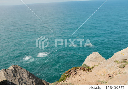 Scenic view of Cabo da Roca steep rocky cliffs meeting blue Atlantic Ocean under bright sky in Sintra Portugal panoramic coastal landscape from westernmost point of mainland Europe 128430271