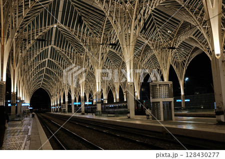 Modern illuminated architecture of Oriente Station at night in Lisbon Portugal featuring geometric roof design arches and empty railway tracks in futuristic urban environment. High quality photo 128430277