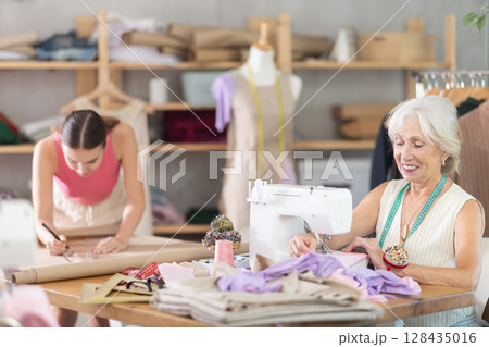 Elderly woman sews on machine female assistant sews on mannequin Elderly woman sews on machine female assistant sews on mannequin 128435016