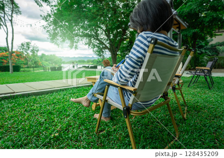 Woman enjoying digital detox while reading book in peaceful garden chair surrounded by green nature for mental wellness and self-reflection time. Social media break. Mindful living and disconnection. 128435209