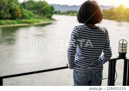 Rear view of a woman gazing at calm river. Travel retreat. Peaceful solitude, personal reflection. Nature connection. Freedom, and emotional healing. Rest, and inner clarity. Emotional balance. 128435210