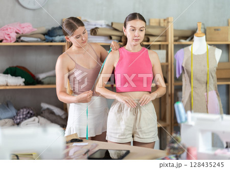 Young seamstress takes body measurements from adult woman Young seamstress takes body measurements from adult woman 128435245