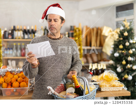 Focused man in festive hat and casualwear looking at shopping list while moving along aisle between displays and holding cart with 128435338