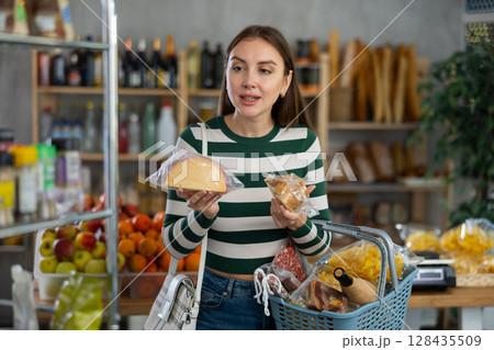 Smiling young girl with shopping basket trying to make choice holding two sorts of cheese in grocery store 128435509