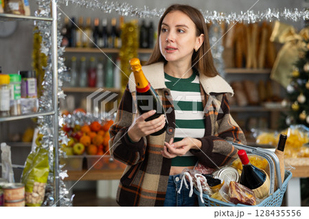 Interested young girl shopper holding bottle and choosing wine for holiday table in grocery hypermarket 128435556