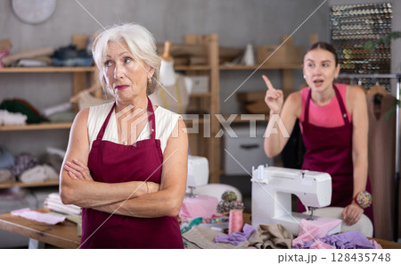 Adult woman scolding elderly woman in sewing workshop Adult woman scolding elderly woman in sewing workshop 128435748