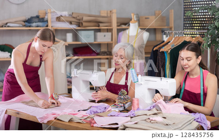Two craftsperson sew clothes, girl assistant attaches pattern to fabric. Two craftsperson sew clothes, girl assistant attaches pattern to fabric. 128435756