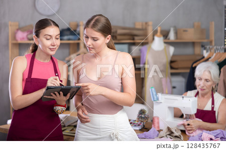 Senior seamstress sewing, apprentice with tablet accept order and talking with customer 128435767