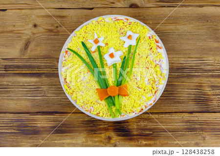 Traditional russian layered salad Mimosa with spring decoration Daffodil on wooden table. Top view. Decoration is made of egg, green onion and carrot 128438258