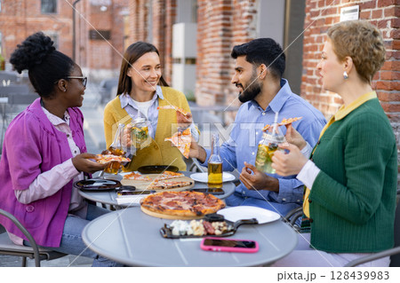 A diverse group of friends enjoys pizza and drinks at an outdoor restaurant, laughing and conversing around a table. 128439983