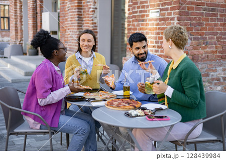 Friends enjoying pizza and drinks together at an outdoor cafe, with smiles and conversation. 128439984