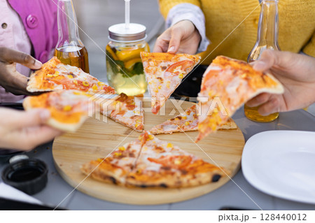 Close-up shot of a group sharing pizza slices, with drinks and a wooden board on the table. 128440012
