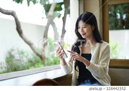 Young woman in cozy casual outfit using mobile phone near a bright cafe window 128442352