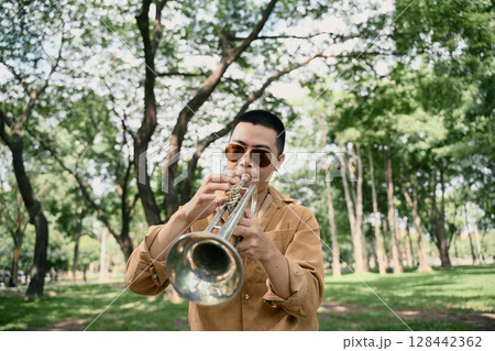 Stylish male artist performing trumpet in a natural setting with trees in the background Stylish male artist performing trumpet in a natural setting with trees in the background 128442362