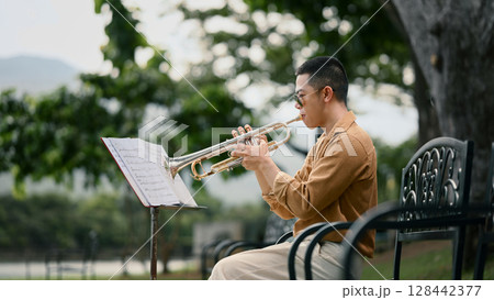 Young man playing trumpet outdoors while reading sheet music in a peaceful park 128442377