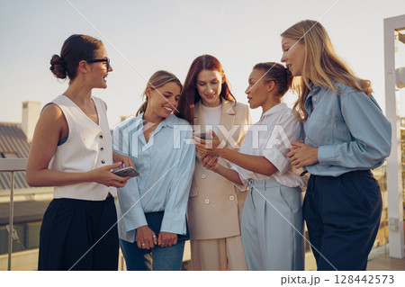 A group of women joyfully engaging in a candid conversation over their smartphones while outdoors, 128442573