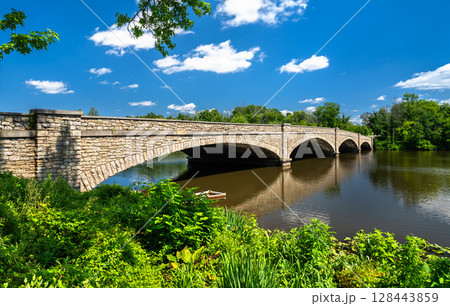 Stone-arched Washington Road Bridge spanning Carnegie Lake in Princeton, New Jersey, United States. The historic structure is surrounded by lush greenery and reflected in the calm lake water beneath a 128443859