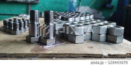 Close-up of new metal bolts and nuts. Steel bolts and nuts. Industrial fasteners displayed on a wooden surface, with a blurred workshop background. Flanged steel hex nuts. Mechanical engineering. 128443870
