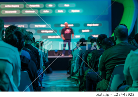 Speaker addressing audience with bright blue conference stage in background 128445922