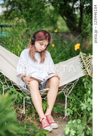 Teenage girl reading and listening to music in hammock outdoors 128446184