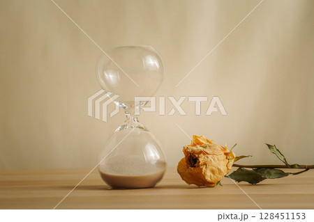 A simple hourglass on a wooden table with dry rose a beige backdrop, symbolizing time flow, aging, and minimalistic living. Represents timeless themes in a creative presentation for stylish concepts. 128451153