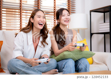 Two asian woman playing video games together with controllers in a cozy living room at home. 128452133