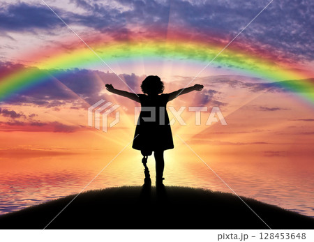 A happy child is a disabled girl with a prosthetic leg in the open air on the background of sea sunset and rainbow 128453648