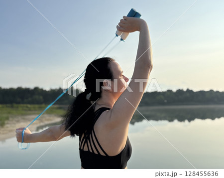 Athletic woman in black sportswear holding a blue skipping rope, standing in nature against the scenic backdrop of a forest and lake. Close up view emphasizing healthy lifestyle, outdoor fitness Athletic woman in black sportswear holding a blue skipping rope, standing in nature against the scenic backdrop of a forest and lake. Close up view emphasizing healthy lifestyle, outdoor fitness 128455636