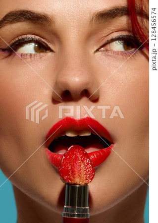 Closeup of woman lips painted red with strawberry on lipstick tube against teal background 128456574