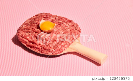 Minced meat and raw egg on ping pong paddle against striped pink background Minced meat and raw egg on ping pong paddle against striped pink background 128456576